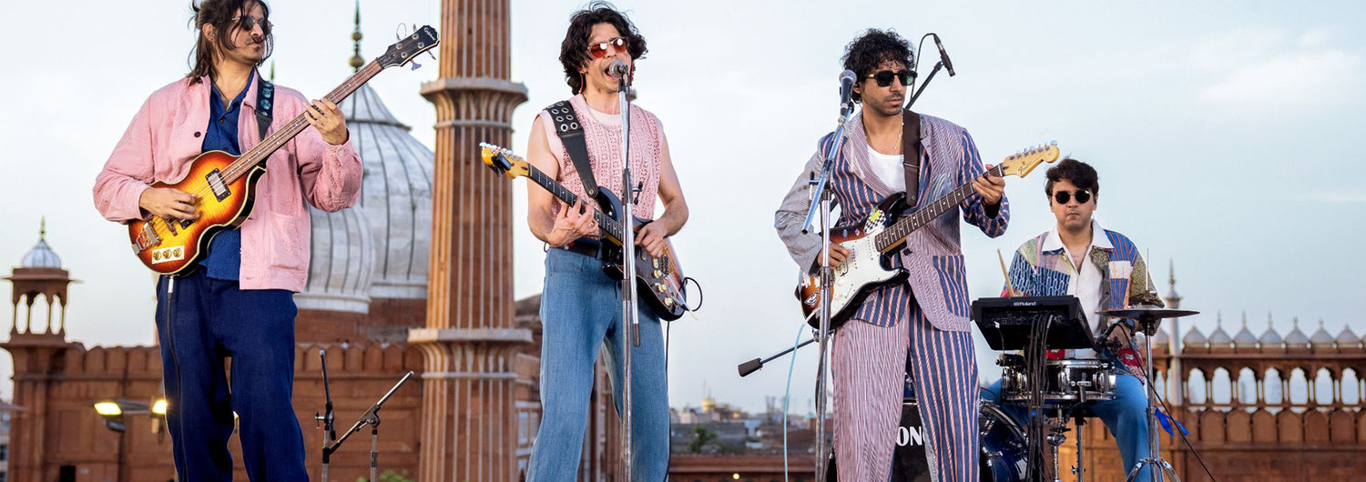 Eine Band spielt auf einem Dach, im Hintergrund sind andere Gebäude zu sehen. Die vier Männer tragen farblich abgestimmte Outfits in den Farben rosa und blau von Kardo. 